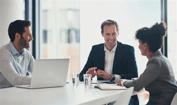 Two men and a woman sitting in an office around a white desk with a laptop, glasses of water and notebooks on it