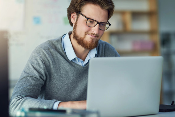 A young man with a beard and glasses smiles gently while working on a laptop at a desk. He wears a gray sweater over a light blue collared shirt. The background features a blurred modern workspace with shelves and written notes, creating a collaborative atmosphere.