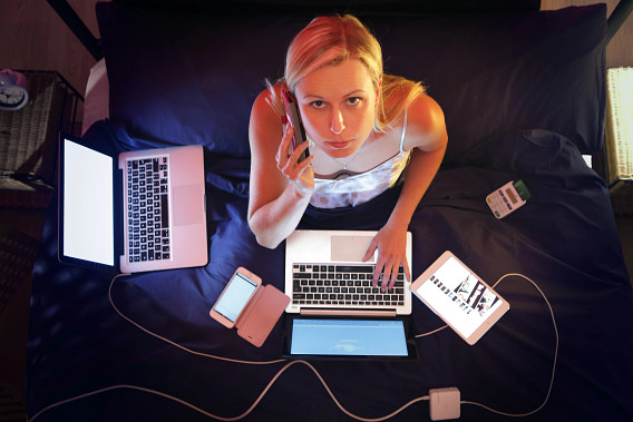 A young woman sitting on a bed, looking up while speaking on a phone. She is surrounded by multiple electronic devices, including a laptop with a blank screen, a tablet displaying images, a smartphone, and a portable charger, all on a dark bedspread. The scene captures a moment of multitasking in a cozy environment.