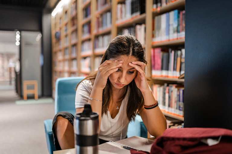 Focused student studying in a library, seated at a table with open books and notes, holding head in hands; background features bookshelves filled with books and a stainless steel water bottle on the table.