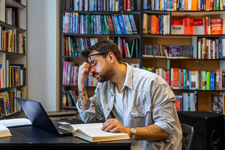 A man wearing glasses and a light denim shirt sits at a desk in a library, looking at a laptop with a pensive expression, his hand resting on his forehead. Open books are spread on the desk, and shelves filled with colorful books surround the space.