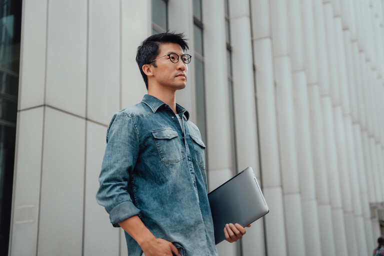 young professional person standing in front of building, holding a laptop