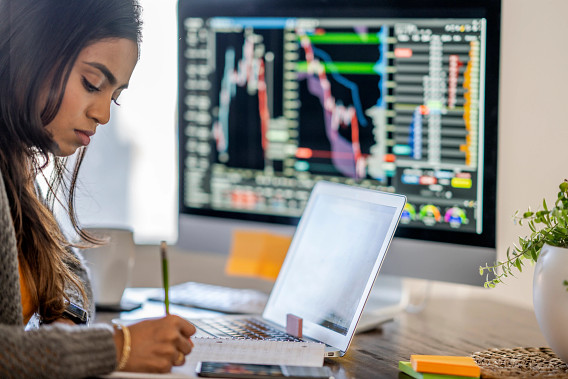 A woman is focused on writing notes at her desk, with a laptop and a large monitor displaying financial charts and data behind her. The setting suggests a work environment involving analysis, trading, or financial planning. A potted plant and stationery add a touch of personalization to the workspace.