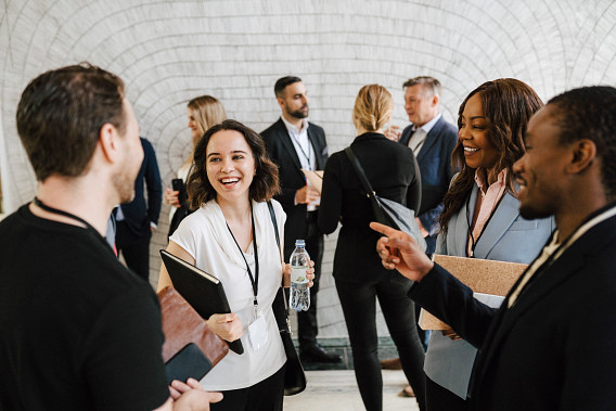 A group of professionals is gathered in a networking event. In the foreground, three people are smiling and conversing, holding notebooks and a water bottle. In the background, others are engaged in discussions. The setting appears formal yet friendly, with an emphasis on collaboration and connection.