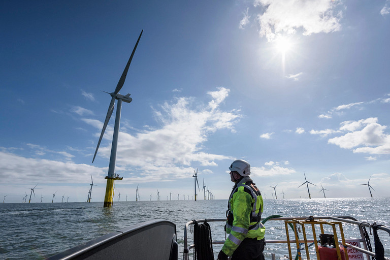 A worker in high-visibility clothing and a helmet stands on a boat, observing a large offshore wind farm. Numerous wind turbines stretch across the horizon under a partly cloudy sky, with sunlight breaking through. The scene highlights renewable energy and marine industry activity.