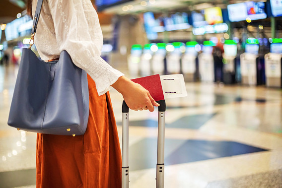 A person at an airport holding a passport and boarding pass, standing next to a suitcase. They are wearing a white blouse and orange skirt, with a blue handbag on their shoulder.